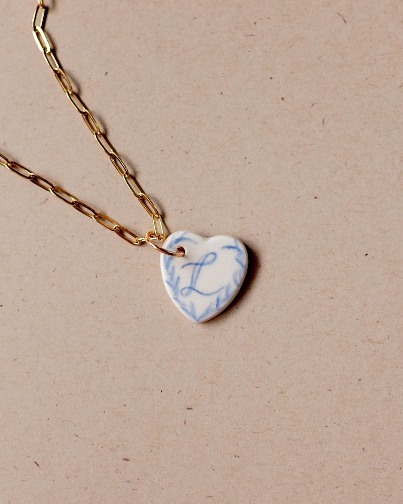 May include: A gold chain necklace with a white ceramic heart pendant. The heart has a blue floral design and the letter "L" in the center.