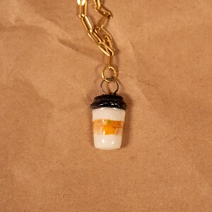Op de afbeelding: Een goudkleurige ketting met een hanger in de vorm van een koffiekopje. De beker is wit met oranje en bruine strepen en een zwart deksel. De ketting wordt getoond tegen een bruine papieren achtergrond.