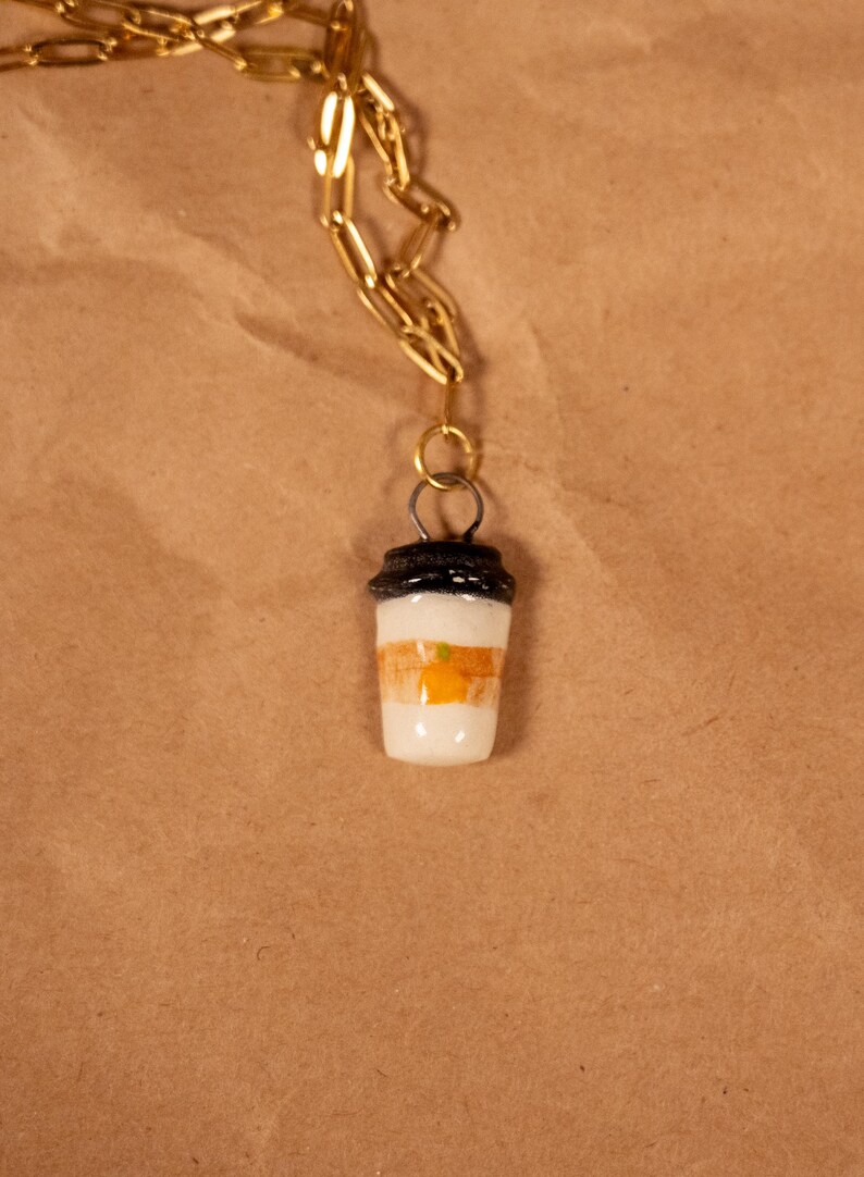 Op de afbeelding: Een goudkleurige ketting met een hanger in de vorm van een koffiekopje. De beker is wit met oranje en bruine strepen en een zwart deksel. De ketting wordt getoond tegen een bruine papieren achtergrond.