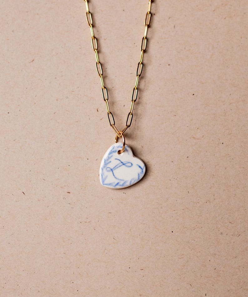 May include: A gold chain necklace with a white porcelain heart pendant. The heart has a blue floral design and the letter "L" in the center.