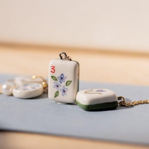 May include: A collection of ceramic pendants, including a rectangular pendant with the number "3" and floral design, and a square pendant with a green border. The pendants are on a gold chain and a pearl necklace. The pendants are white with blue and green accents.