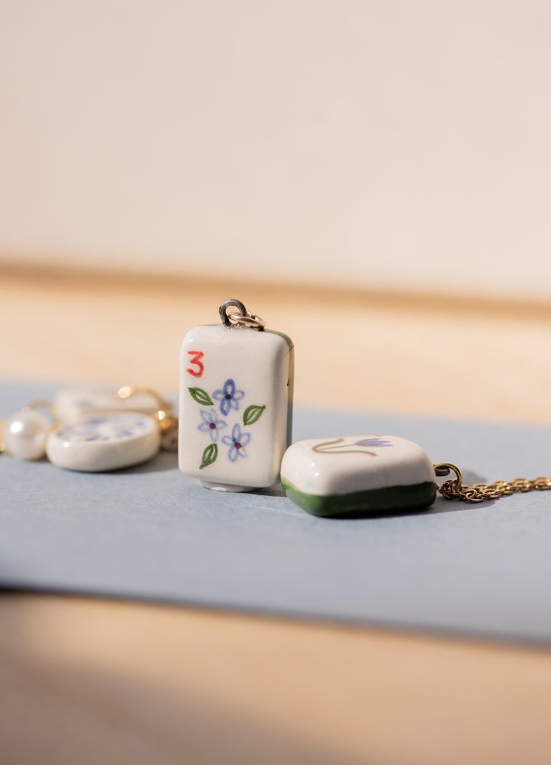 May include: A collection of ceramic pendants, including a rectangular pendant with the number "3" and floral design, and a square pendant with a green border. The pendants are on a gold chain and a pearl necklace. The pendants are white with blue and green accents.