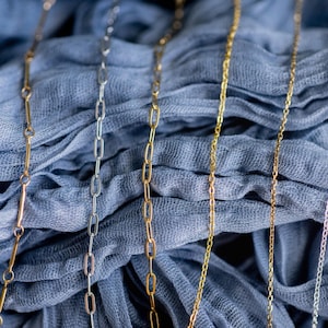May include: Close-up of several delicate necklaces in gold, silver, and rose gold tones. The chains have different link styles, including paperclip and delicate chain designs. The necklaces are displayed on a draped blue fabric.