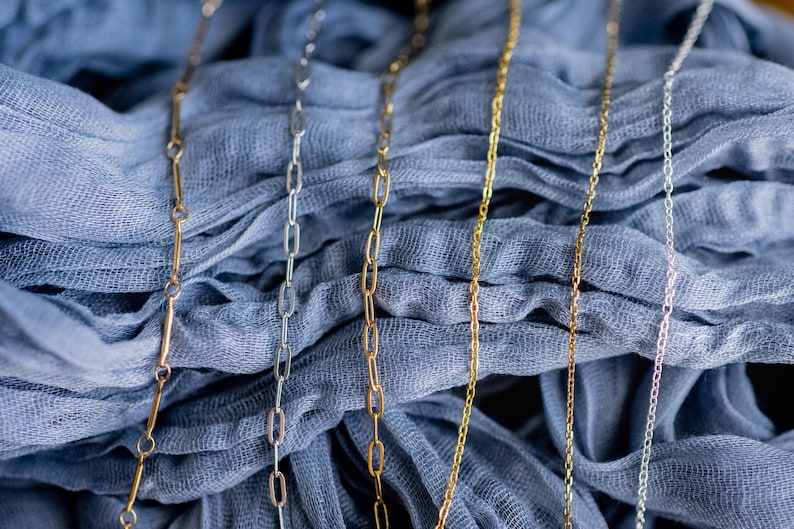 May include: Close-up of several delicate necklaces in gold, silver, and rose gold tones. The chains have different link styles, including paperclip and delicate chain designs. The necklaces are displayed on a draped blue fabric.