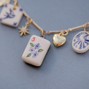 May include: A gold-toned necklace with multiple charms. Charms include a heart, a pearl, a square with a bird, a star, and a rectangular tile with the number 3 and floral details. The charms are white with blue accents.