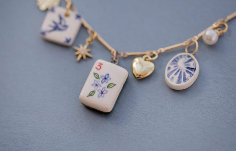 May include: A gold-toned necklace with multiple charms. Charms include a heart, a pearl, a square with a bird, a star, and a rectangular tile with the number 3 and floral details. The charms are white with blue accents.