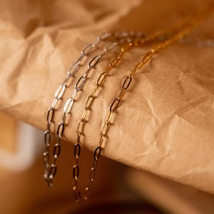 Op de afbeelding: Close-up van drie delicate kettingkettingen in zilver- en goudtinten. De kettingen hebben een ovaal schakelontwerp en worden gepresenteerd op een getextureerd bruin oppervlak. De kettingen zijn een eenvoudig en elegant sieraad.