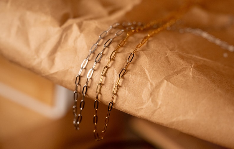Op de afbeelding: Close-up van drie delicate kettingkettingen in zilver- en goudtinten. De kettingen hebben een ovaal schakelontwerp en worden gepresenteerd op een getextureerd bruin oppervlak. De kettingen zijn een eenvoudig en elegant sieraad.