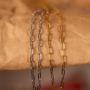 Op de afbeelding: Drie delicate kettingkettingen in zilver- en goudtinten. De kettingen hebben een paperclip-schakels ontwerp, met langwerpige rechthoekige schakels. De kettingen worden gepresenteerd tegen een bruine papieren achtergrond.
