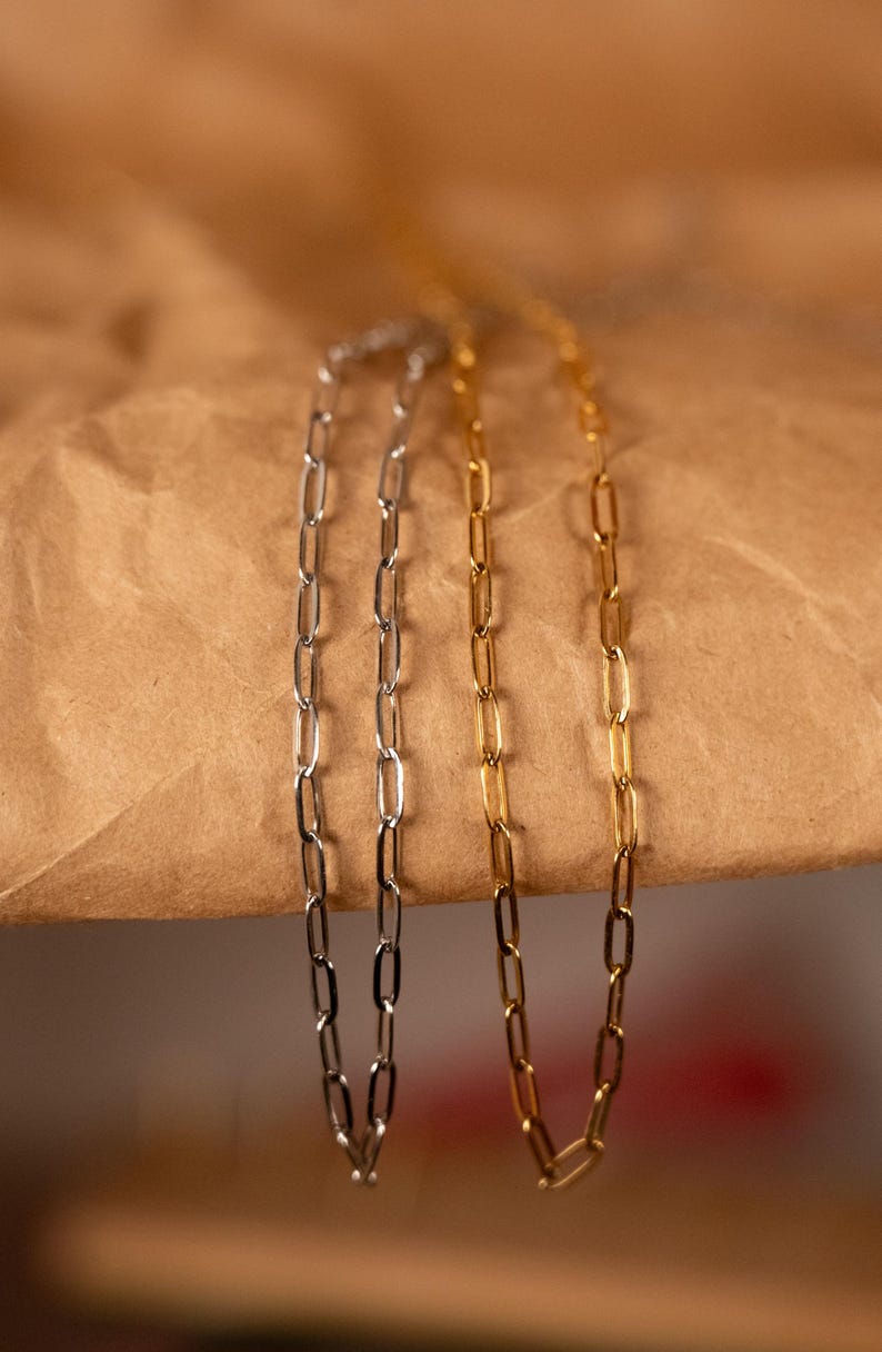 Op de afbeelding: Drie delicate kettingkettingen in zilver- en goudtinten. De kettingen hebben een paperclip-schakels ontwerp, met langwerpige rechthoekige schakels. De kettingen worden gepresenteerd tegen een bruine papieren achtergrond.