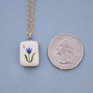 May include: A gold-colored chain necklace with a small, rectangular pendant. The pendant is white with a blue flower design and the number "1". A U.S. quarter coin is next to the pendant.