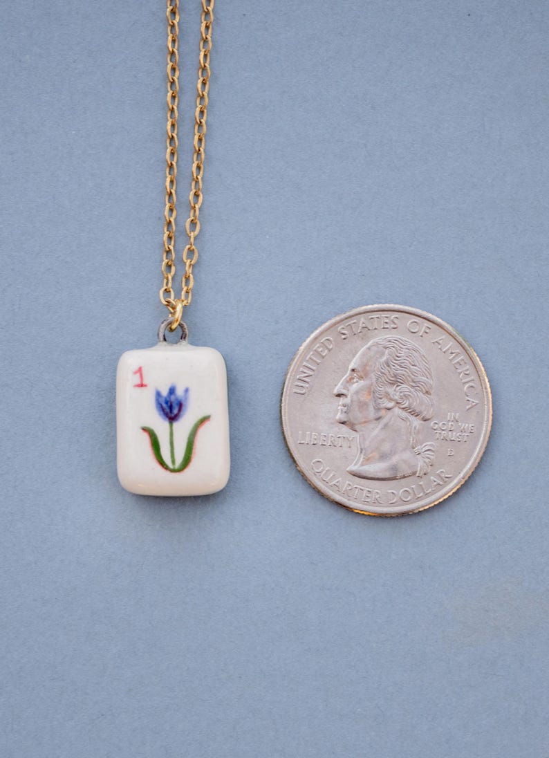 May include: A gold-colored chain necklace with a small, rectangular pendant. The pendant is white with a blue flower design and the number "1". A U.S. quarter coin is next to the pendant.