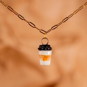 Op de afbeelding: Een gouden ketting met een hanger in de vorm van een koffiebeker. De hanger heeft een zwart deksel, een witte body met oranje strepen en een klein groen accent. Een eenvoudig, elegant accessoire.