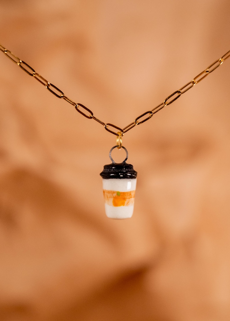 Op de afbeelding: Een gouden ketting met een hanger in de vorm van een koffiebeker. De hanger heeft een zwart deksel, een witte body met oranje strepen en een klein groen accent. Een eenvoudig, elegant accessoire.