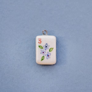 May include: A rectangular white pendant with a silver loop at the top. The pendant features a red number 3, blue flowers, and green leaves. The background is a solid light blue.
