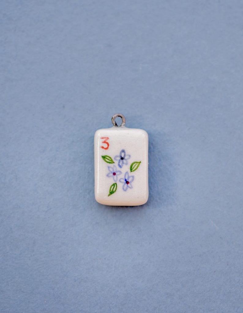 May include: A rectangular white pendant with a silver loop at the top. The pendant features a red number 3, blue flowers, and green leaves. The background is a solid light blue.