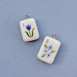 May include: Two white ceramic pendants with silver-colored loops. One pendant has a red "1" and a blue flower with green stem. The other has a red "3" and blue flowers with green leaves. The pendants are rectangular.