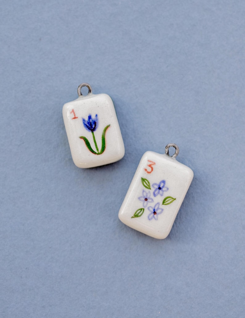 May include: Two white ceramic pendants with silver-colored loops. One pendant has a red "1" and a blue flower with green stem. The other has a red "3" and blue flowers with green leaves. The pendants are rectangular.