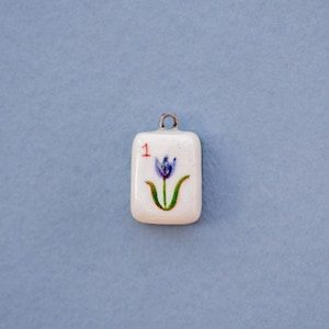 May include: A small, rectangular pendant with a white background and a painted blue tulip design. The number "1" is in red in the upper left corner. The pendant has a silver-colored loop at the top for hanging.
