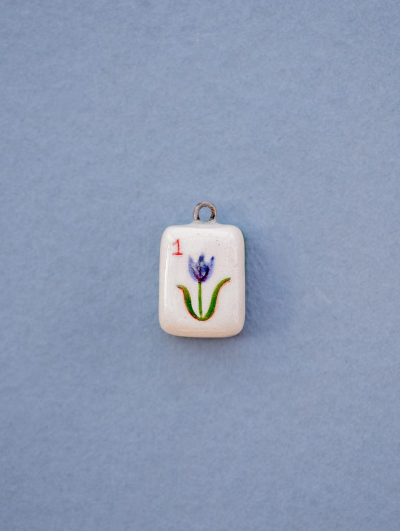 May include: A small, rectangular pendant with a white background and a painted blue tulip design. The number "1" is in red in the upper left corner. The pendant has a silver-colored loop at the top for hanging.
