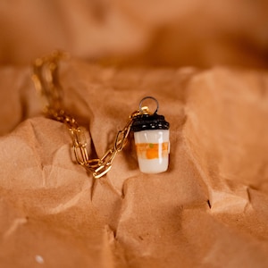 Op de afbeelding: Een goudkleurige ketting met een hanger in de vorm van een koffiebeker. De beker is wit met oranje en zwarte details, liggend op een bruin, getextureerd oppervlak.