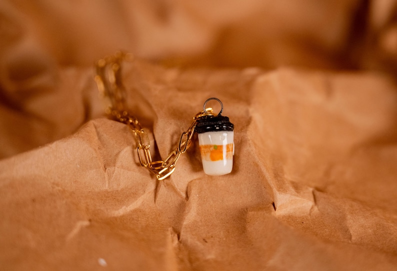 Op de afbeelding: Een goudkleurige ketting met een hanger in de vorm van een koffiebeker. De beker is wit met oranje en zwarte details, liggend op een bruin, getextureerd oppervlak.