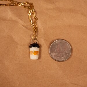 Op de afbeelding: Een goudkleurige ketting met een kleine hanger in de vorm van een koffiebeker. De beker is wit met oranje en zwarte details. Een munt ligt naast de hanger ter vergelijking van de grootte.