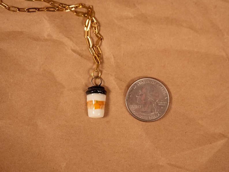Op de afbeelding: Een goudkleurige ketting met een kleine hanger in de vorm van een koffiebeker. De beker is wit met oranje en zwarte details. Een munt ligt naast de hanger ter vergelijking van de grootte.