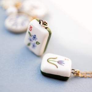May include: Two white ceramic pendant necklaces with gold chains. One pendant features the number "3" and blue floral details. The other pendant has a purple flower design. Both pendants have green edges.