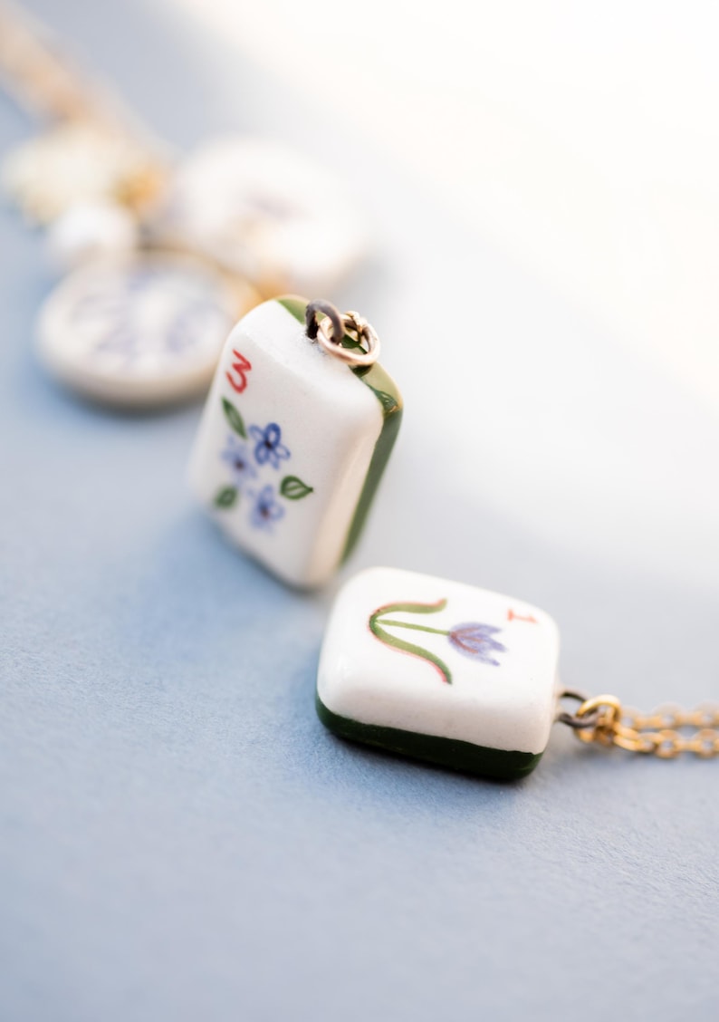 May include: Two white ceramic pendant necklaces with gold chains. One pendant features the number "3" and blue floral details. The other pendant has a purple flower design. Both pendants have green edges.