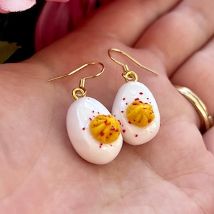May include: A pair of gold dangle earrings shaped like deviled eggs. The earrings are white with a yellow yolk and red paprika.