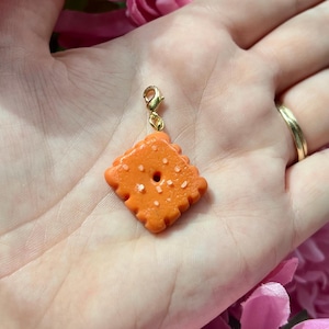 May include: A gold charm shaped like a square cracker with a hole in the center. The cracker is orange with white sprinkles.