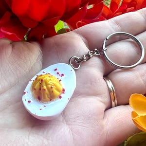 May include: A white and yellow egg-shaped keychain with red sprinkles and a yellow yolk. The keychain is attached to a silver key ring.