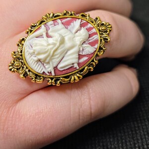 Puede incluir: Un anillo de tono dorado con un camafeo que presenta un &aacute;ngel esculpido en blanco con alas sobre un fondo rojo.