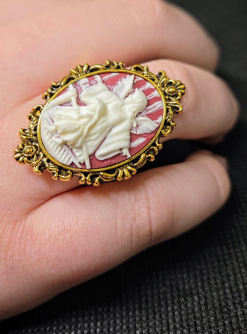 Puede incluir: Un anillo de tono dorado con un camafeo que presenta un &aacute;ngel esculpido en blanco con alas sobre un fondo rojo.