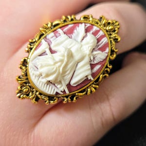 Puede incluir: Un anillo de tono dorado con un camafeo que presenta una figura esculpida en blanco de una mujer sobre un fondo rojo. El anillo tiene un dise&ntilde;o ornamentado con detalles intrincados.