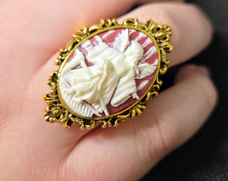 Puede incluir: Un anillo de tono dorado con un camafeo que presenta una figura esculpida en blanco de una mujer sobre un fondo rojo. El anillo tiene un dise&ntilde;o ornamentado con detalles intrincados.