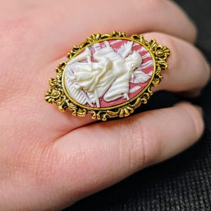Puede incluir: Un anillo de tono dorado con un camafeo que presenta un &aacute;ngel blanco con alas sobre un fondo rojo. El camafeo est&aacute; engastado en un marco ovalado adornado.
