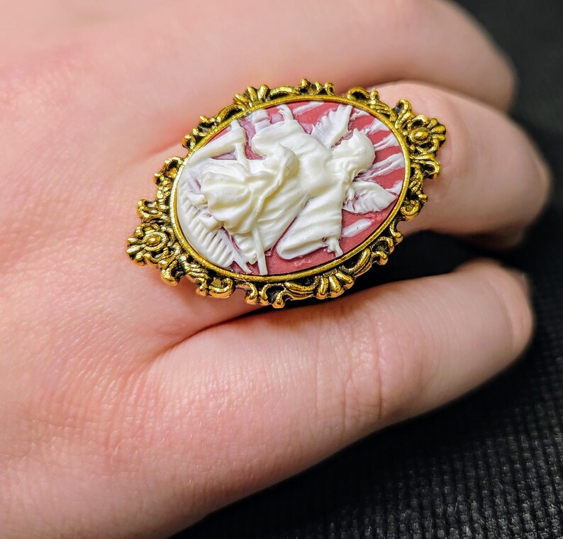 Puede incluir: Un anillo de tono dorado con un camafeo que presenta un &aacute;ngel blanco con alas sobre un fondo rojo. El camafeo est&aacute; engastado en un marco ovalado adornado.