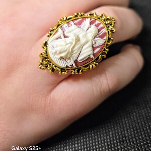 Puede incluir: Un anillo de tono dorado con un camafeo que presenta una figura blanca sobre un fondo rojo. El camafeo est&aacute; rodeado por un marco dorado adornado.