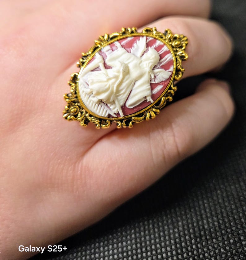 Puede incluir: Un anillo de tono dorado con un camafeo que presenta una figura blanca sobre un fondo rojo. El camafeo est&aacute; rodeado por un marco dorado adornado.