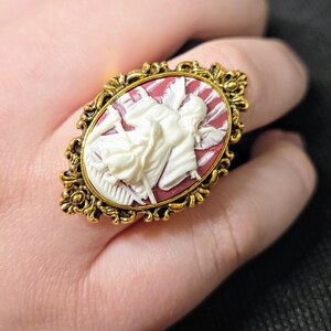 Puede incluir: Un anillo de tono dorado con un camafeo que representa un &aacute;ngel blanco con alas sobre un fondo rojo.