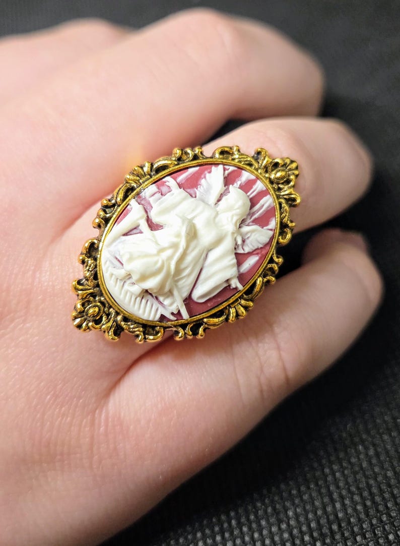 Puede incluir: Un anillo de tono dorado con un camafeo que representa un &aacute;ngel blanco con alas sobre un fondo rojo.