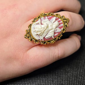Puede incluir: Un anillo de tono dorado con un camafeo que presenta una figura blanca en un fondo rojo y blanco. El camafeo est&aacute; engastado en un marco ovalado con detalles ornamentados.