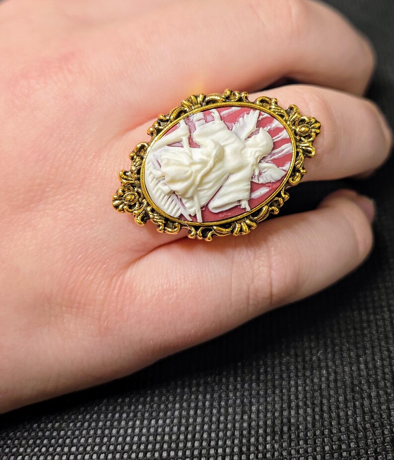 Puede incluir: Un anillo de tono dorado con un camafeo que presenta una figura blanca en un fondo rojo y blanco. El camafeo est&aacute; engastado en un marco ovalado con detalles ornamentados.