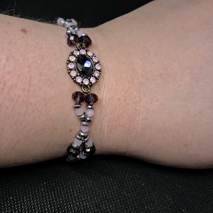 May include: A beaded bracelet with a central oval-shaped dark purple gem surrounded by smaller pink gems. The bracelet features alternating sections of pink and silver beads, and dark purple beads.