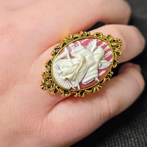 Puede incluir: Un anillo de tono dorado con un camafeo que presenta una figura blanca sobre un fondo rojo. El camafeo est&aacute; engastado en un marco ovalado con detalles ornamentados.