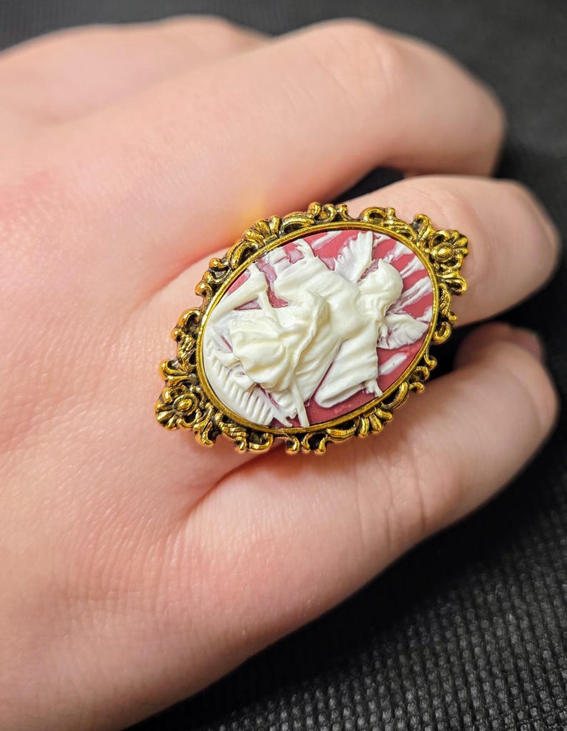 Puede incluir: Un anillo de tono dorado con un camafeo que presenta una figura blanca sobre un fondo rojo. El camafeo est&aacute; engastado en un marco ovalado con detalles ornamentados.