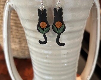 May include: Black cat silhouette earrings with a small orange flower and green leaves. The earrings are hanging from a white ceramic vase.