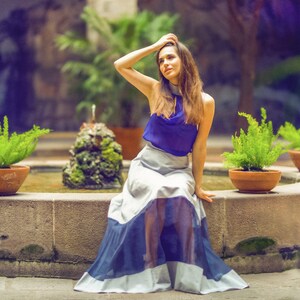 May include: A woman wearing a blue top and a white and blue skirt sits on a stone ledge by a fountain. She has long brown hair and is looking to the side. The fountain is surrounded by greenery.